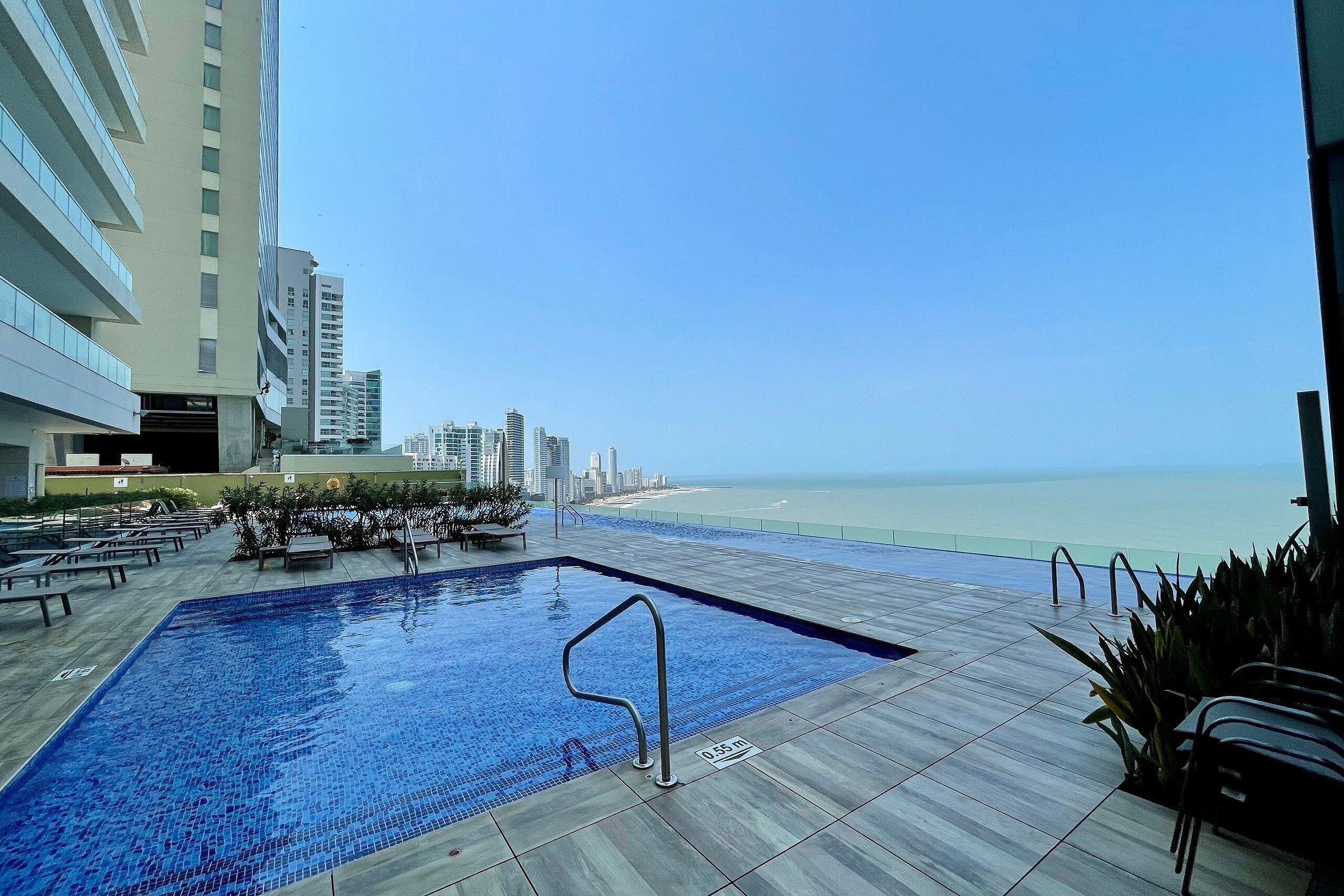 Une piscine avec un escalier en métal, entourée d’une terrasse carrelée, offrant une vue sur une plage et le paysage urbain.
