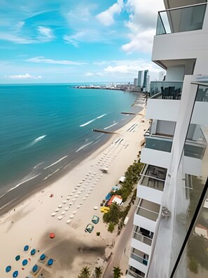 Une vue sur la plage depuis un immeuble de grande hauteur, avec un ciel dégagé, une plage de sable bordée de parasols et une jetée visible au loin.