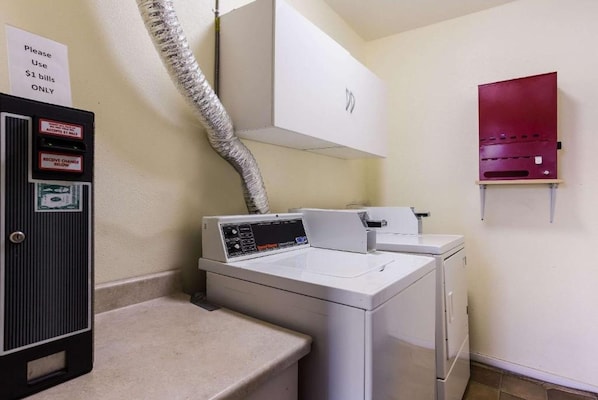 A laundry area with a washing machine, dryer, and a utility cabinet with a sign instructing to use $1 bills only. - Urban Inn & Suites (Boise)