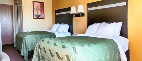 A hotel room with two beds, each with a green bedspread and white pillows. There is a wall-mounted lamp above each bed.