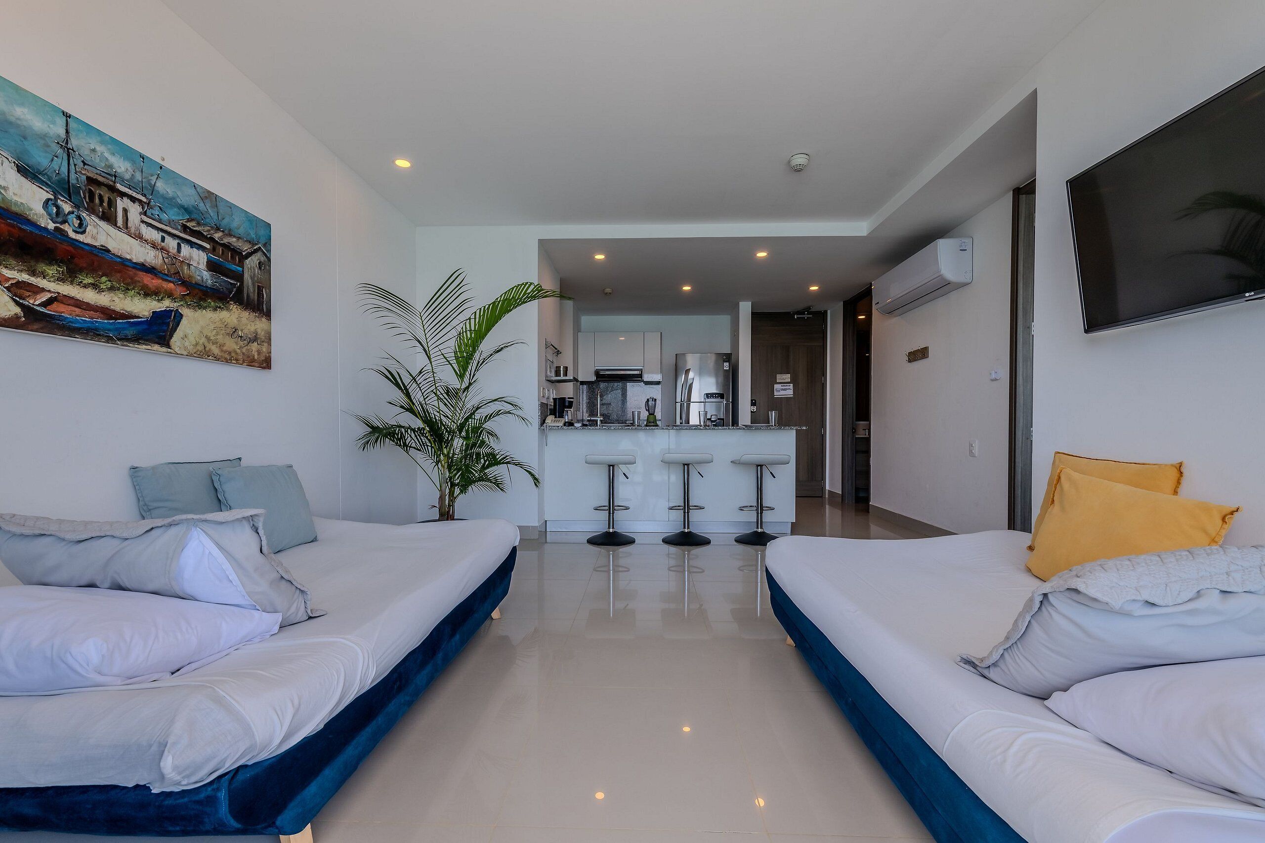 Habitación de hotel con dos camas, una zona de cocina y un televisor montado en la pared.
