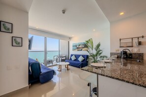 A spacious living area with a large window, a blue sofa, a small round table, and a kitchen area with granite countertops. - 1704 Morros City - Spectacular Apt in Bocagrande (Cartagena)