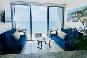 Ein Wohnzimmer mit zwei blauen Sofas, einem Couchtisch und einer großen Schiebeglasdoor, die auf einen Balkon mit Meerblick führt.