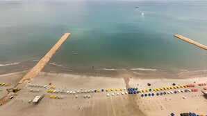 Ein Luftbild zeigt einen Strand mit einem hölzernen Steg, der ins Meer führt, einen sandigen Uferbereich mit bunten Bojen und ein ruhiges Meer.