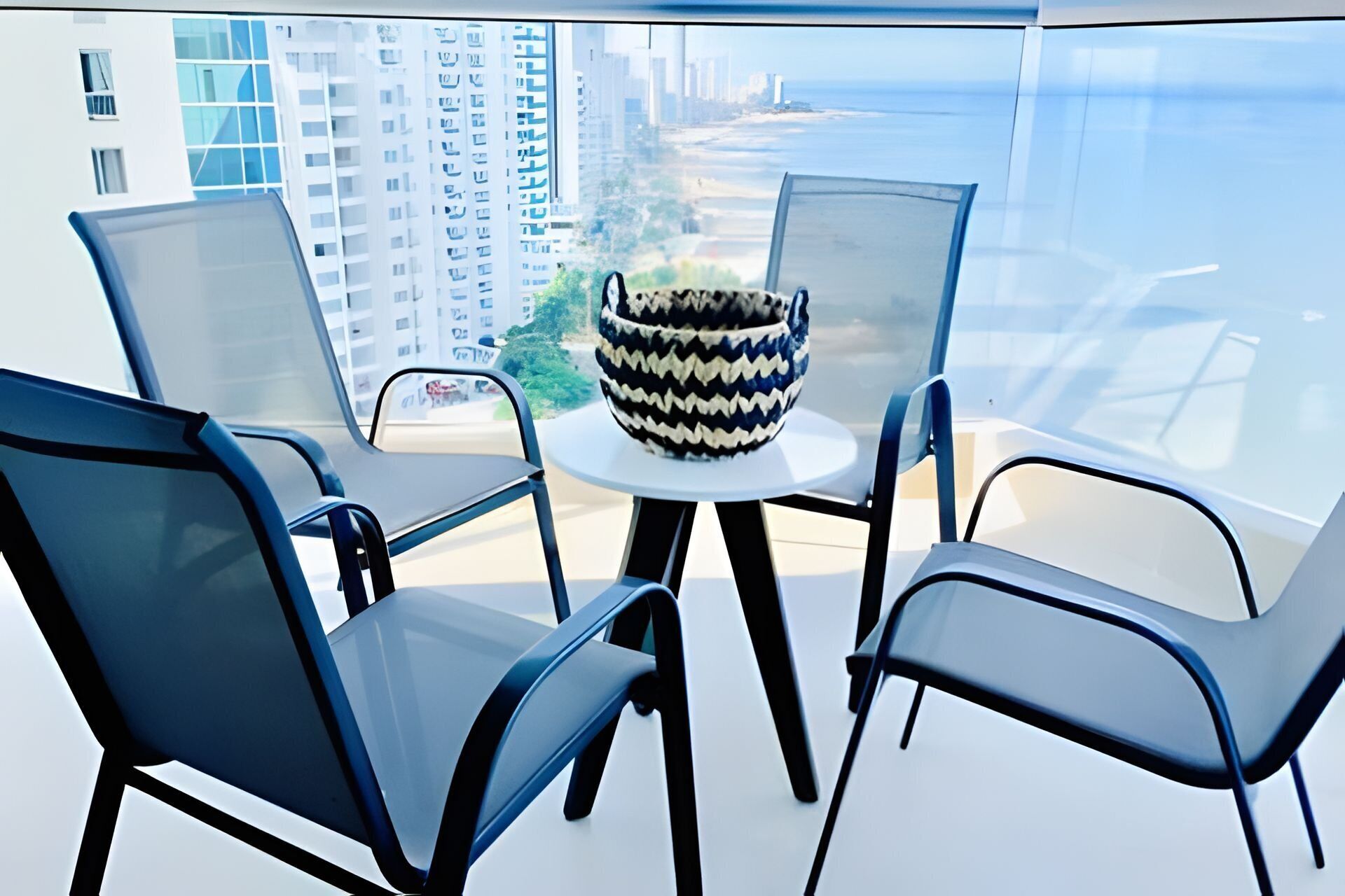 Une chambre moderne avec une table et des chaises en verre, un vase décoratif et une vue sur le paysage urbain et la plage.