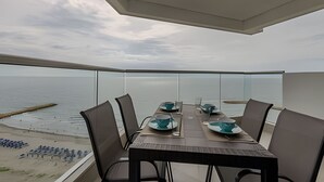 Ein Essbereich mit einem Tisch für vier Personen, mit Blick auf einen Strand und ruhiges Wasser unter einem bewölkten Himmel.