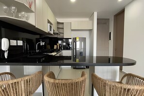 A modern kitchen with a black countertop, stainless steel appliances, wicker chairs, and a door in the background. - Infinitum Spectacular 23rd floor 1 BR Condo (Cartagena)