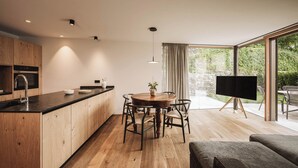 A modern kitchen with wooden cabinets, a black countertop, and a dining area with a round wooden table and chairs. There is a large window with a view of greenery outside. - Garden Apartment Bachquell (Brixen)
