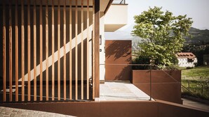A modern building with a wooden slatted entrance, a concrete pathway, and a view of a tree and distant hills. - Garden Apartment Bachquell (Brixen)