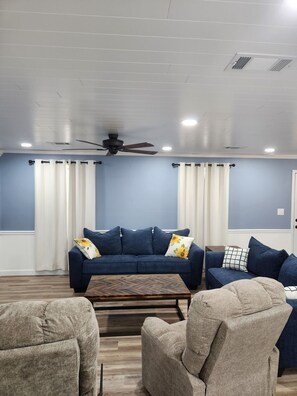 A living room with a blue sofa, two armchairs, a wooden coffee table, and a ceiling fan. - Renovated Lakefront Home with Covered Porch, Pool & Playground (Hot Springs)
