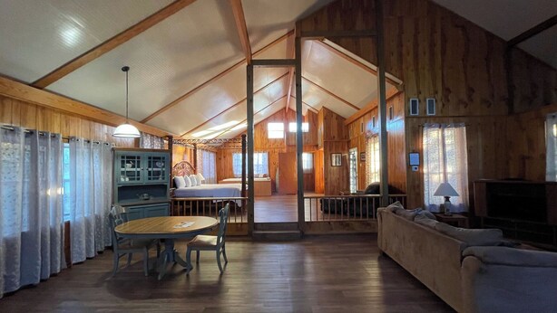 A spacious loft-style room with a high ceiling, wooden walls, a dining area with a table and chairs, a sofa, and a television. - Couples Retreat with King Canopy Bed, Jacuzzi for Two, Fireplace & Pond-View Porch (Zwolle)