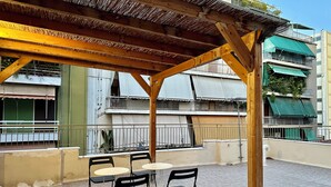 En udendørs terrasse med et træbeklædt pergola, sorte metalstole og et lille rundt bord.