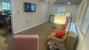 A room with a ceiling fan, a television mounted on the wall, a patterned ottoman, a sofa with a blanket, and a window with blinds.