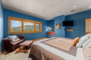 A bedroom with a large bed, a wooden bench, a red leather sofa, a dresser, a TV, and a window with a mountain view.