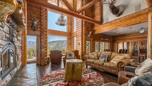 A log cabin interior with a stone fireplace, wooden walls, a round rug, and a large window overlooking a mountainous landscape. - Miracle Lodge - Hot Tub, Golf & Mountain Views (Leavenworth)