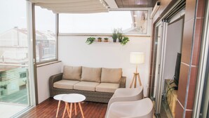 Un balcon avec un canapé en osier, deux chaises blanches, une petite table ronde et un lampadaire.