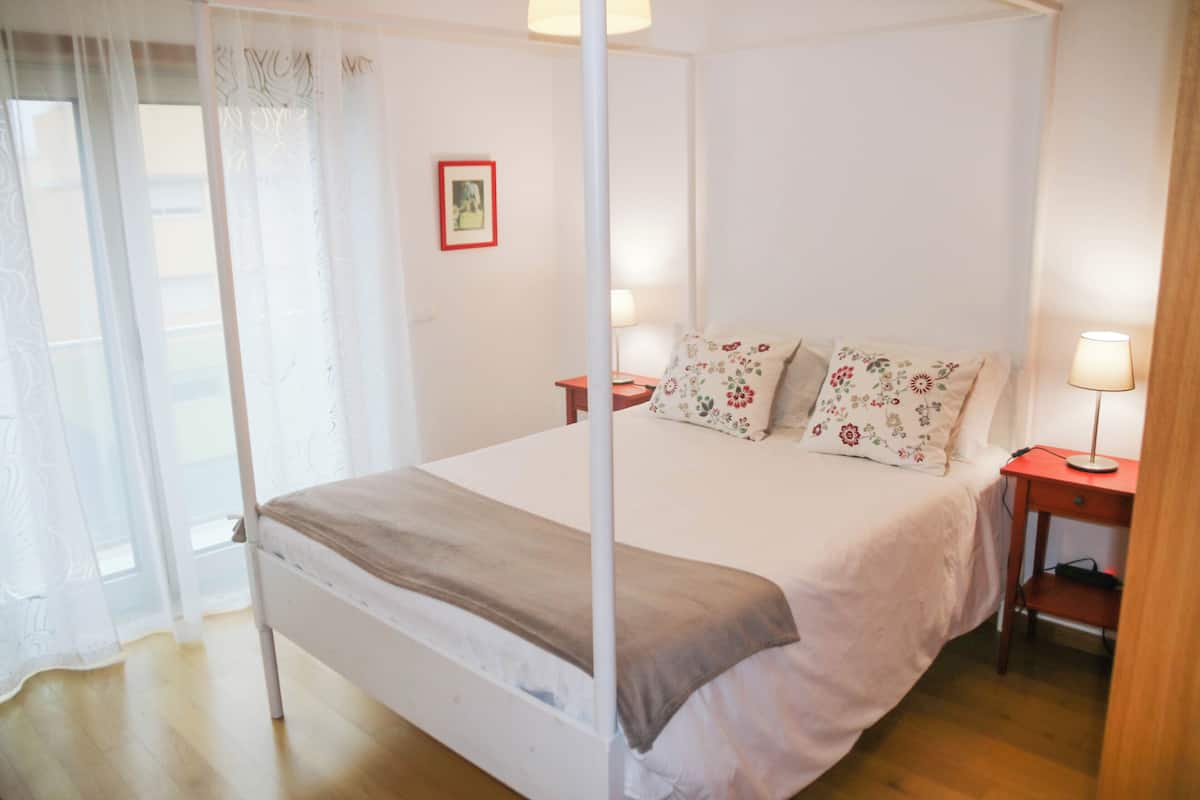 A four-poster bed with a white canopy, two bedside tables with lamps, floral-patterned pillows, and a framed picture on the wall.