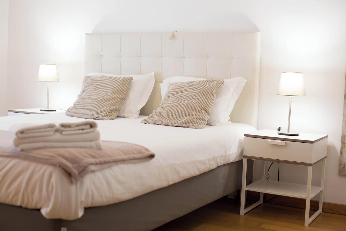 A neatly made bed with two pillows, a bedside table with a lamp, and a small shelf.