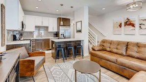 A modern kitchen with white cabinets, a central island with bar stools, a leather sofa, and a staircase leading to another level. - Trekker Townhome | Mountain Charm | New Floors! (Georgetown)
