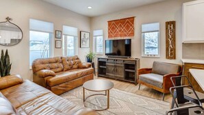 A modern living room with a leather sofa, a coffee table, a TV, and a kitchen area with a countertop and cabinets. - Trekker Townhome | Mountain Charm | New Floors! (Georgetown)