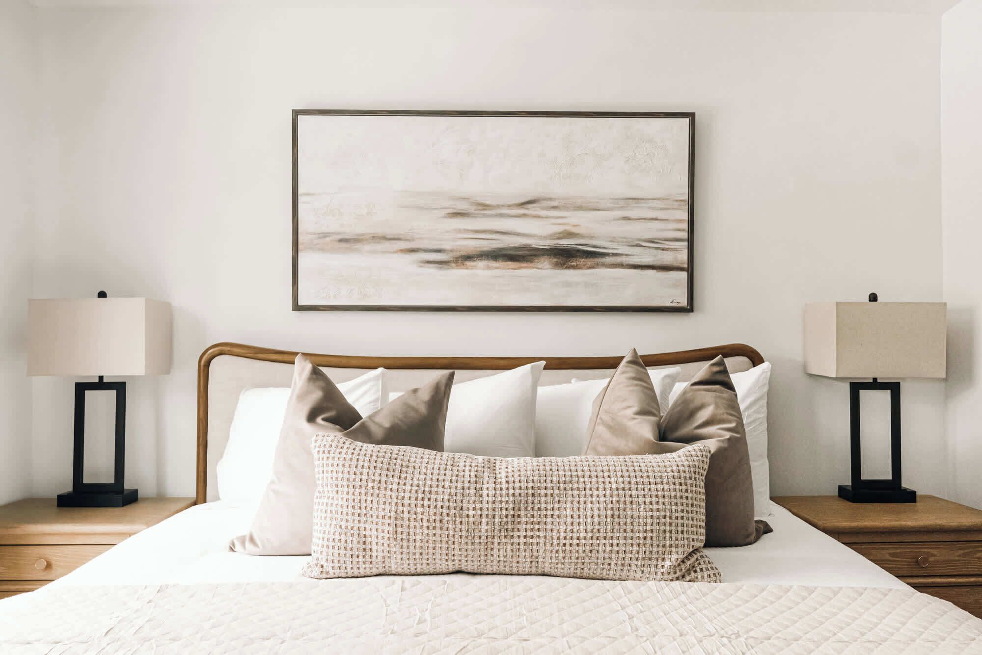 A neatly made bed with a quilted white duvet, a wooden headboard, and several pillows in neutral tones. There is a framed abstract painting above the bed, and two bedside lamps on wooden nightstands.