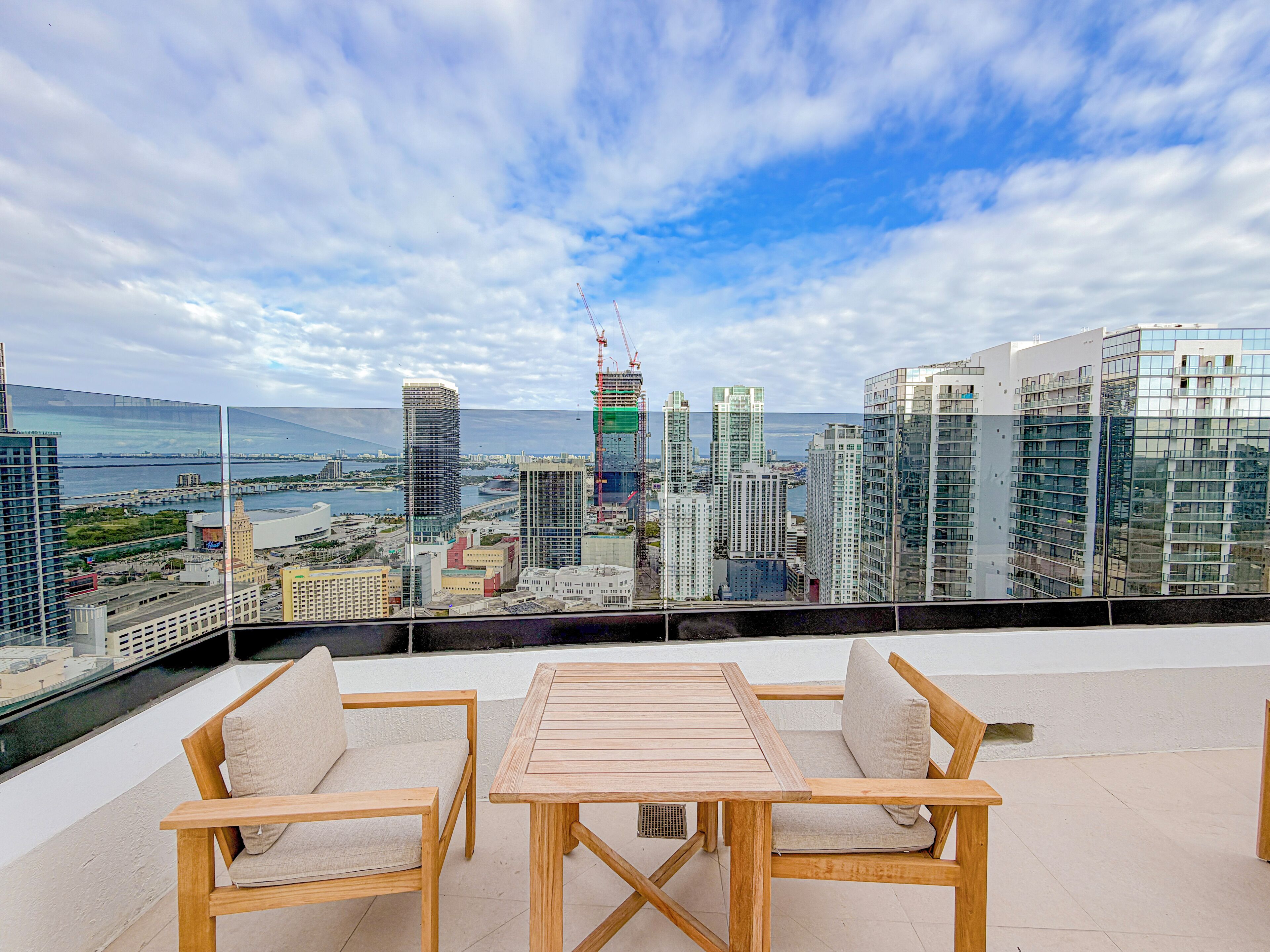 Une terrasse sur le toit, avec une table en bois et deux chaises, d’où l’on peut admirer un panorama sur la ville, avec ses bâtiments modernes et une vue sur la mer.
