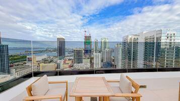 Une terrasse sur le toit, avec une table en bois et deux chaises, d’où l’on peut admirer un panorama sur la ville, avec ses bâtiments modernes et une vue sur la mer.