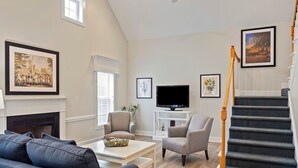 Ein geräumiges Wohnzimmer mit Kamin, Fernseher, Treppe und Deckenventilator.