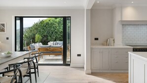 A modern kitchen with white cabinets, a built-in oven, and a dining area with a wooden table and chairs. - Luxury seaside home | 5 mins from beach | sea view | hot tub (Downderry)