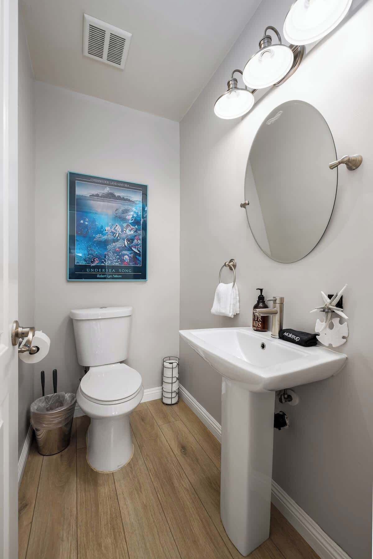 Een moderne badkamer met toilet, wastafel en een grote spiegel. Er hangt een poster aan de muur en een prullenbak is zichtbaar.