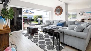 Ein modernes Wohnzimmer mit einem grauen Sofa, einem Couchtisch und einem gemusterten Teppich. Ein großes Fenster bietet Blick ins Freie.