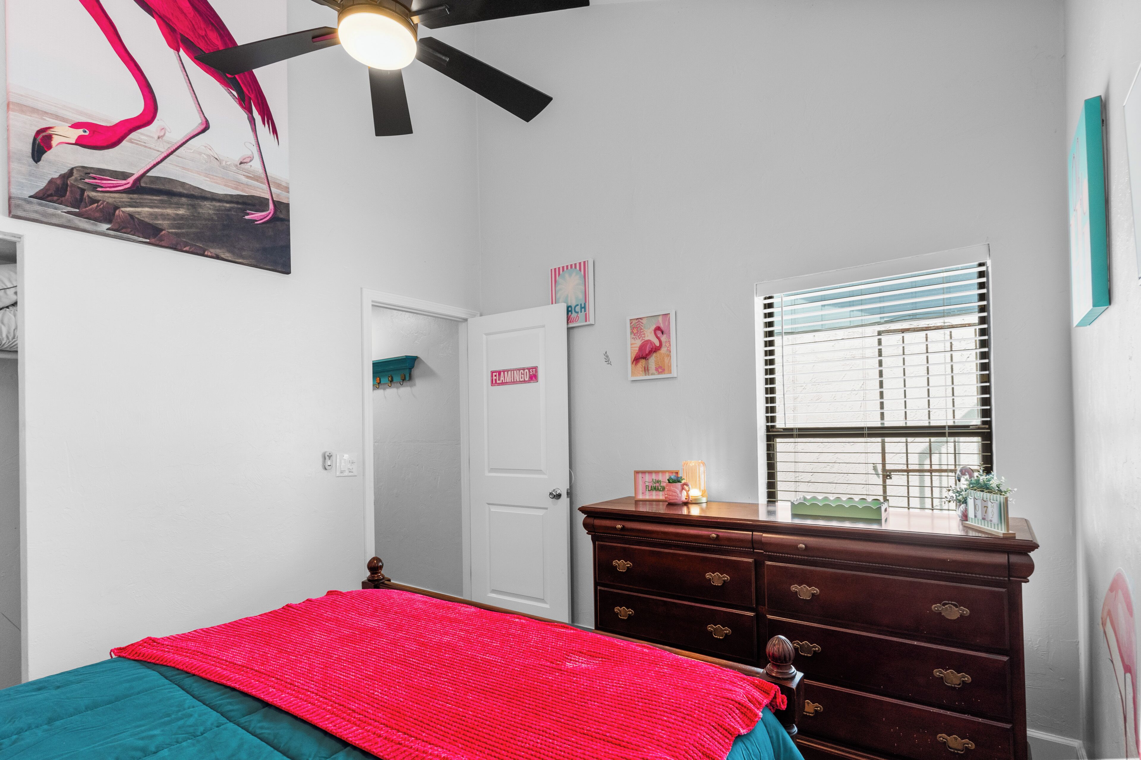 Ein Schlafzimmer mit Bett, Kommode, Deckenventilator, einem Fenster mit Jalousien sowie Wanddekorationen, darunter ein Flamingo-Gemälde.