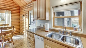 Interieur van een blokhut met een houten eettafel, een ingebouwde vaatwasser, een dubbele spoelbak en een raam met uitzicht op een witte picket fence.