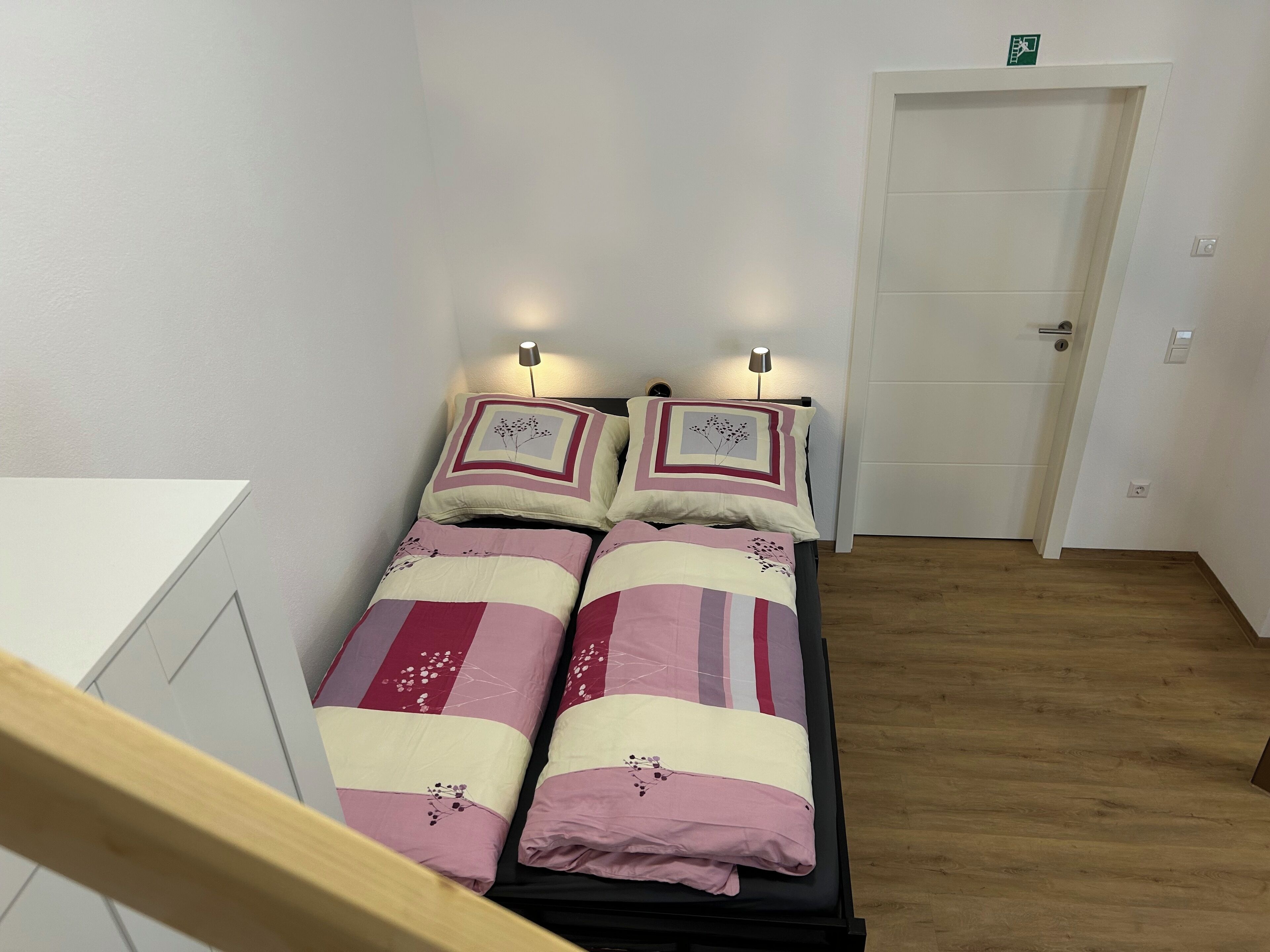 A twin bed with a floral-patterned duvet, two bedside lamps, and a white door in a room with wooden flooring.
