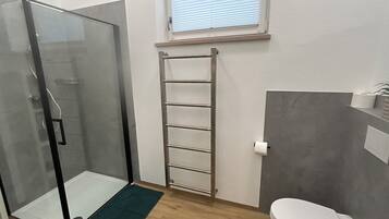 A bathroom with a glass shower enclosure, a wooden floor, a green mat, a towel rack, and a toilet.