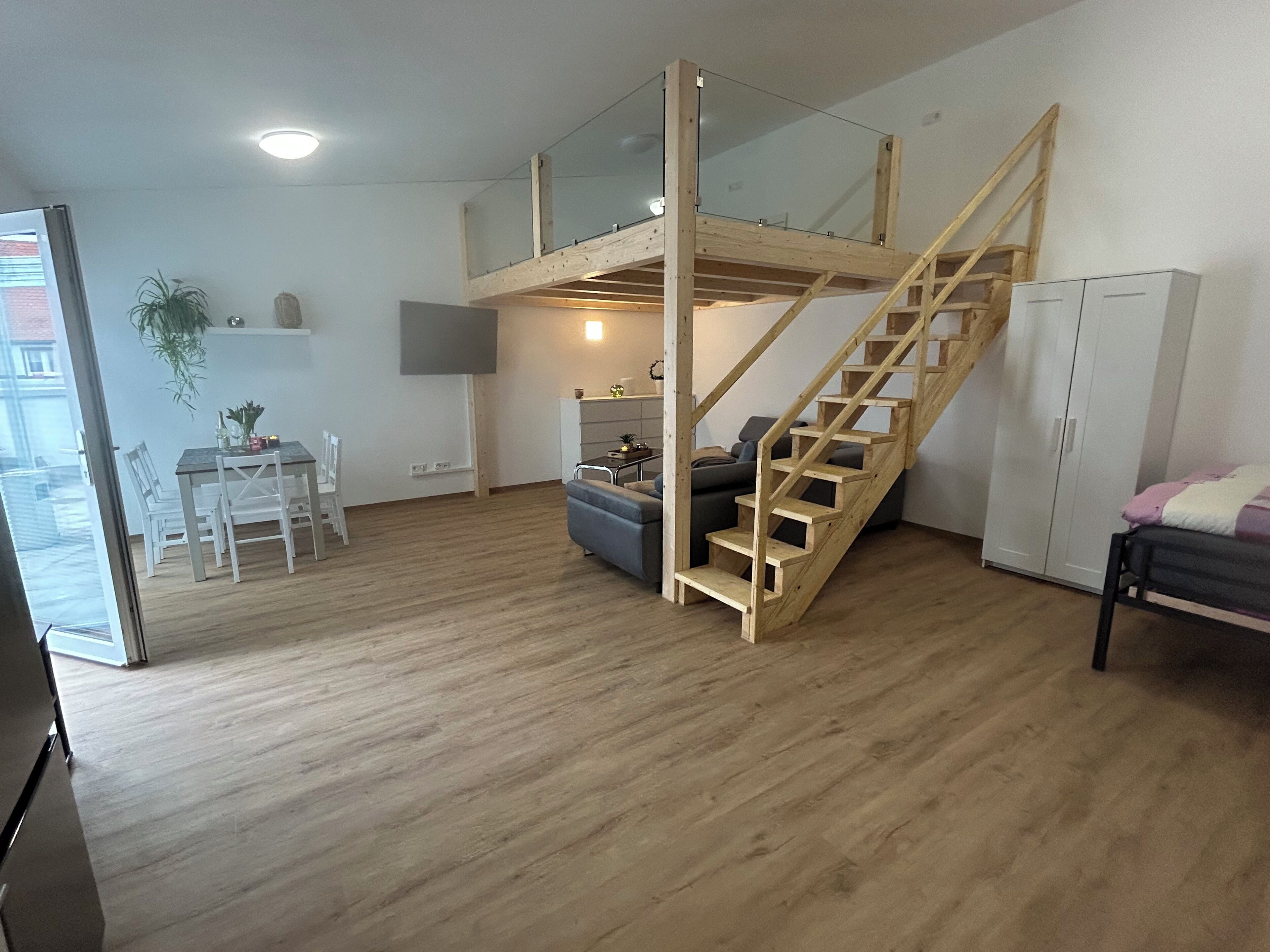 Een ruime woonkamer met een houten trap naar een hoger gelegen verdieping, een eettafel met stoelen, een bank en een kast.