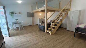Een ruime woonkamer met een houten trap naar een hoger gelegen verdieping, een eettafel met stoelen, een bank en een kast.