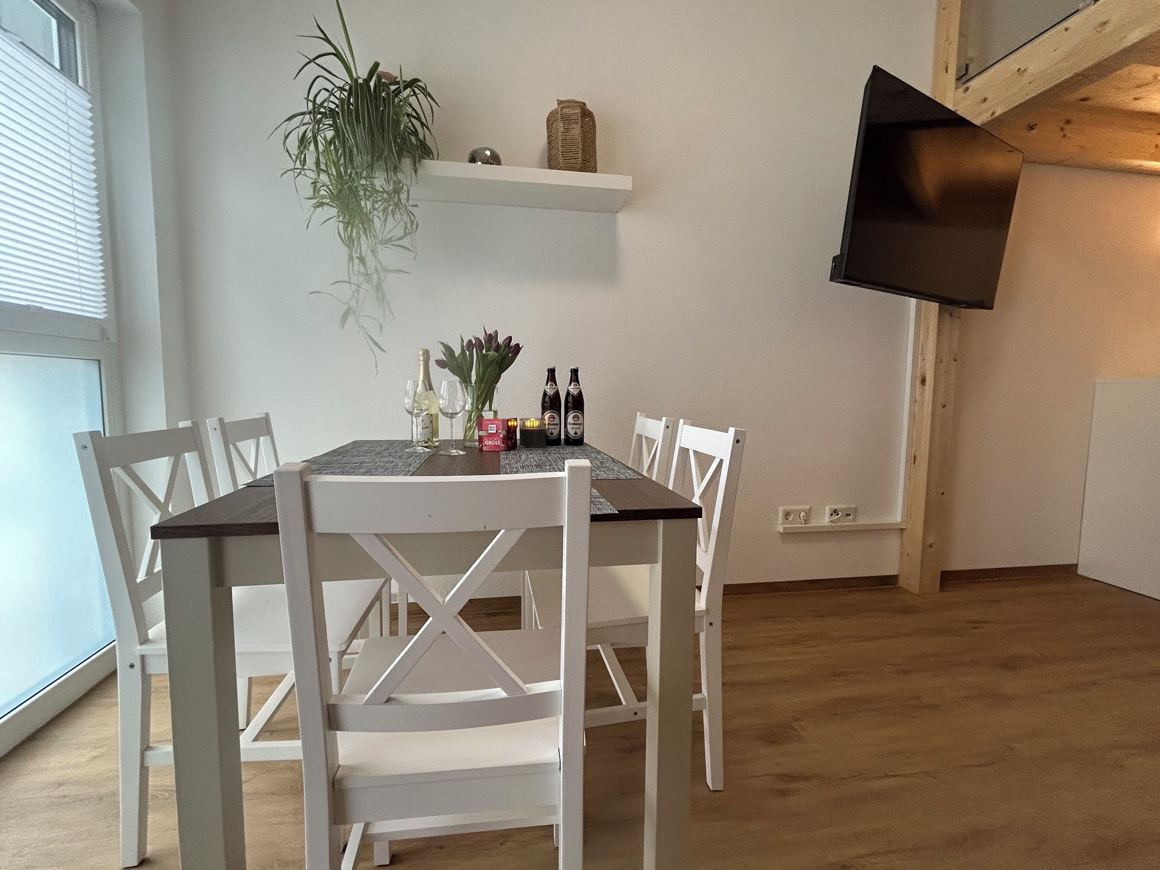 A dining area with a table set for four, surrounded by white chairs. A plant hangs on the wall, and a shelf above holds a basket and decorative items.