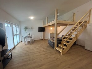 A spacious living area with a wooden staircase, a dining table with chairs, a sofa, and a TV mounted on the wall. - Beautiful new 1-room apartment ideal for hikers (Ederheim)