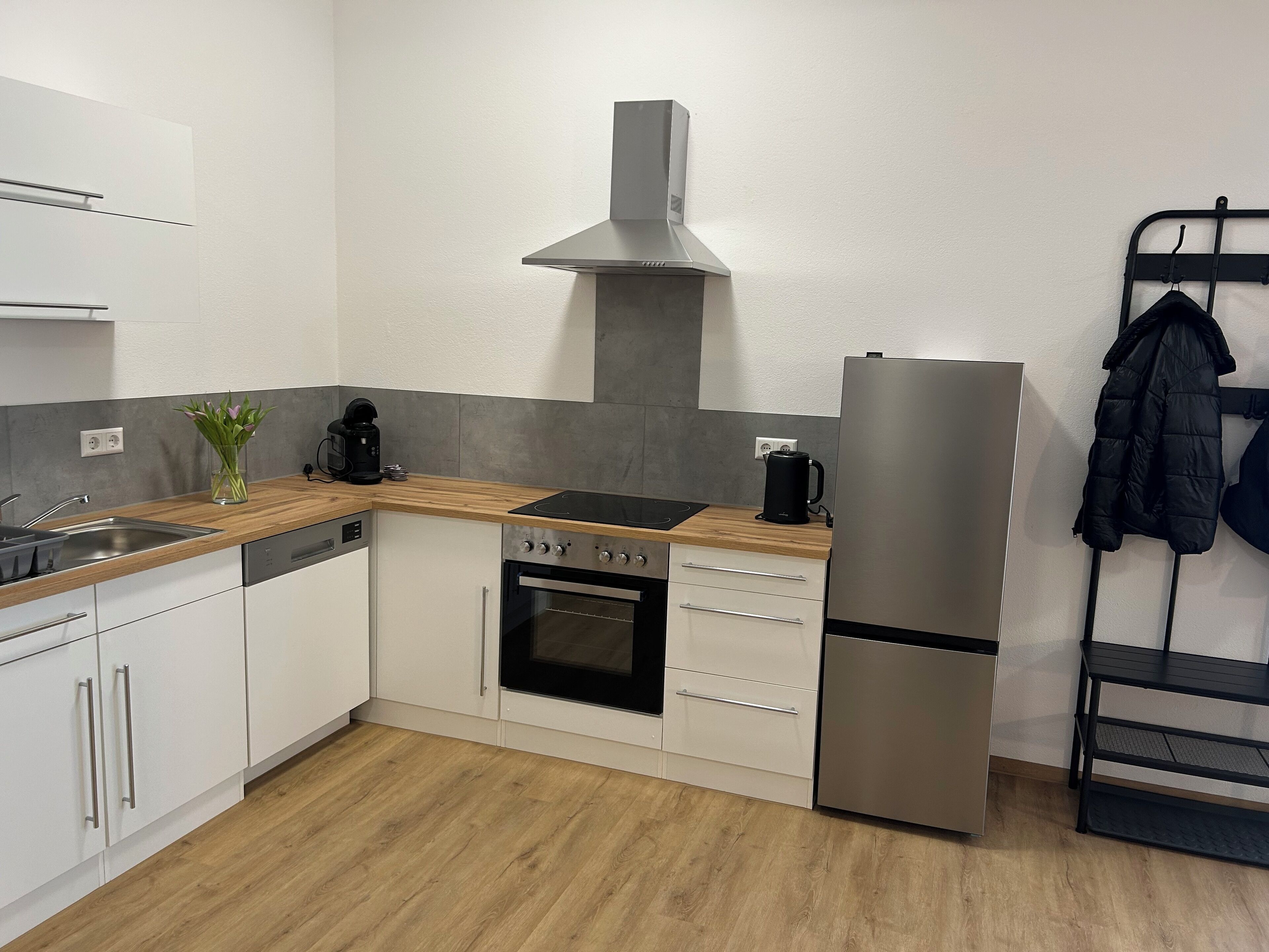 A modern kitchen with white cabinets, a stainless steel refrigerator, a built-in oven, a coffee maker, and a hanging rack with a jacket.