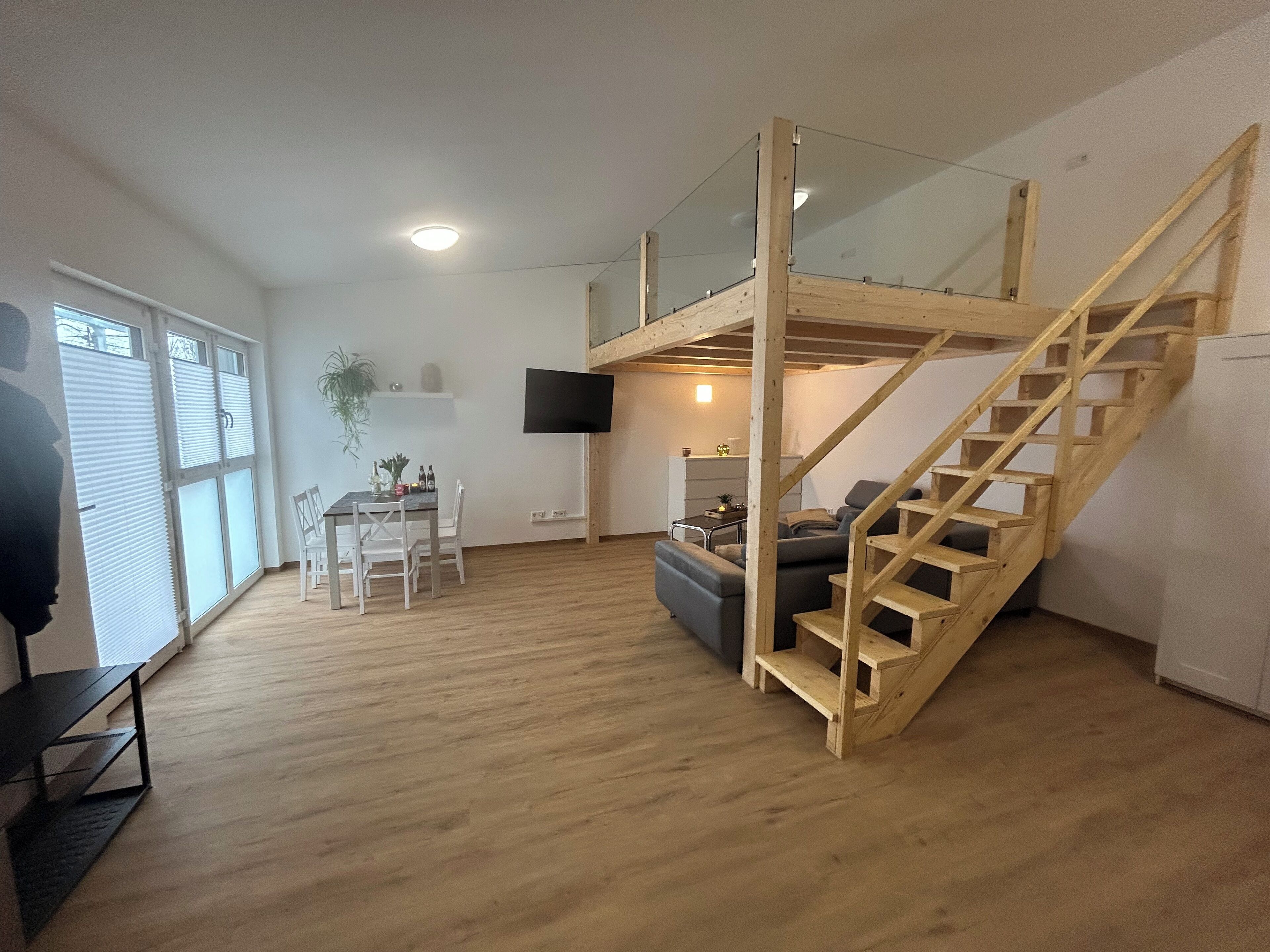 A spacious living area with a wooden staircase, a dining table with chairs, a sofa, and a flat-screen TV mounted on the wall.
