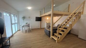 A spacious living area with a wooden staircase, a dining table with chairs, a sofa, and a flat-screen TV mounted on the wall.