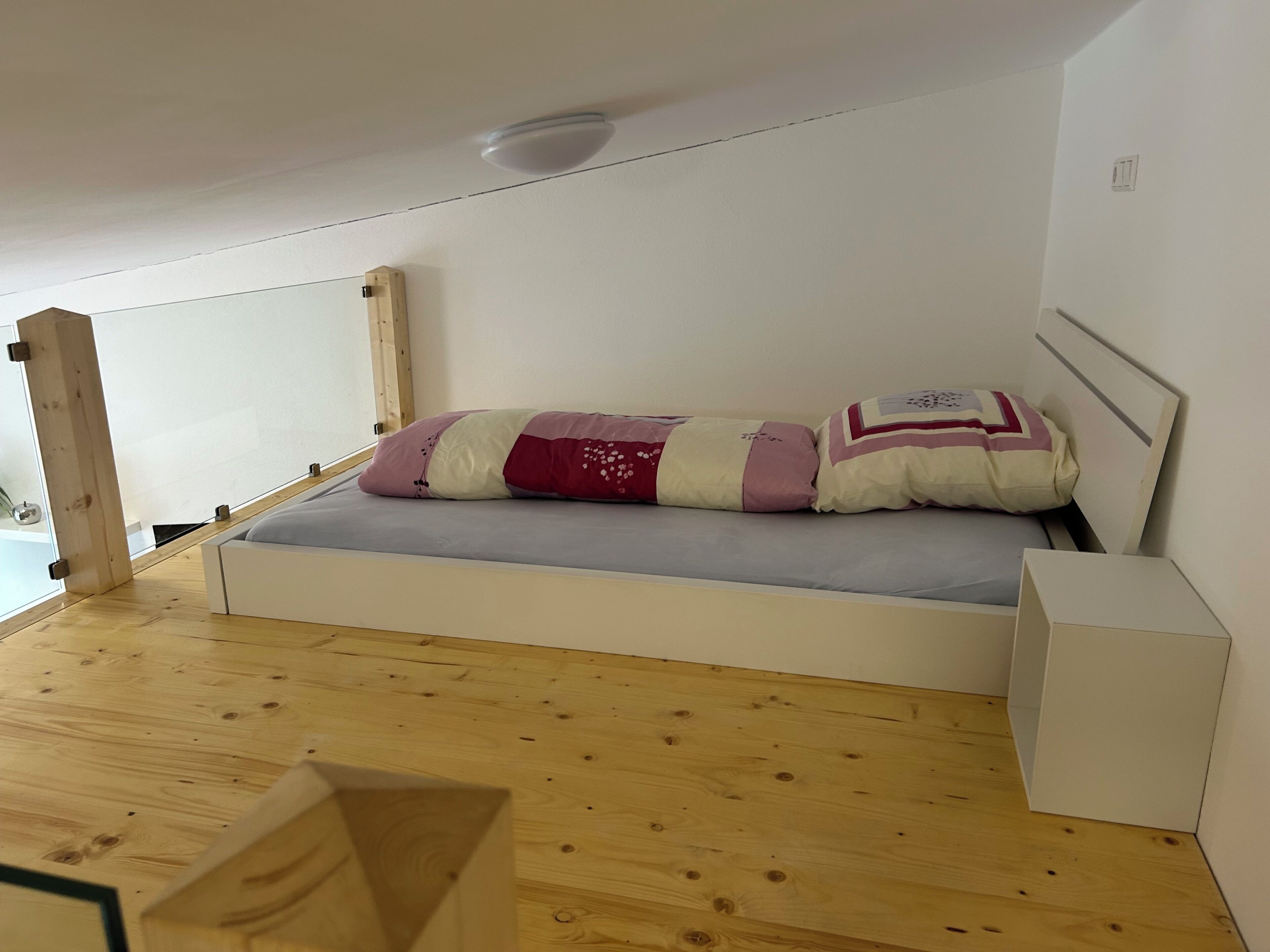 A small, single-bed room with a wooden floor, white walls, and a simple bed with a patterned blanket.