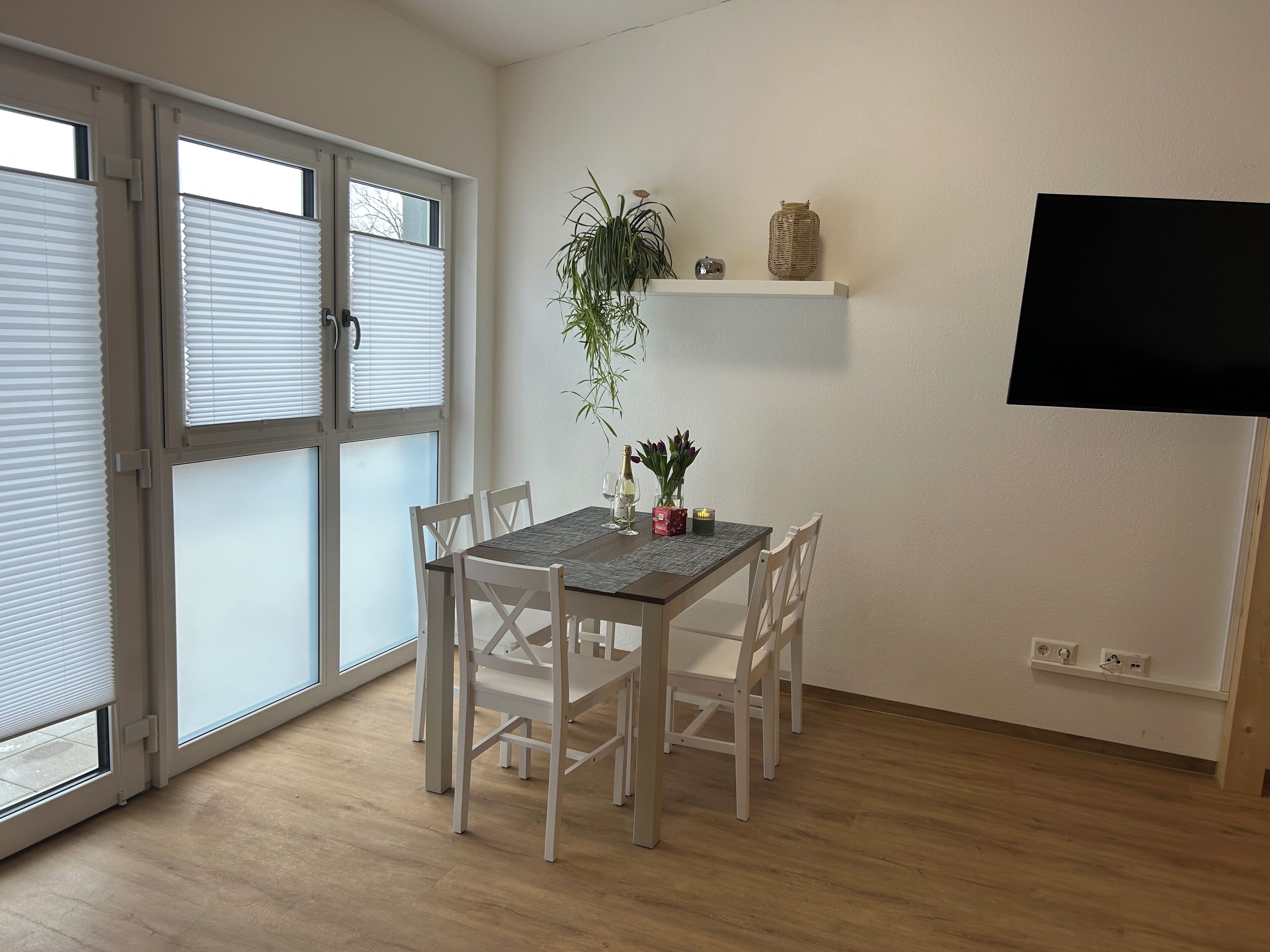 A dining area with a table set for two, white chairs, a potted plant, and a shelf with decorative items.