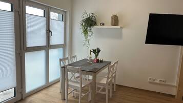 A dining area with a table set for two, white chairs, a potted plant, and a shelf with decorative items.
