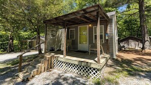 A rustic cabin with a porch, a wooden staircase, and a small shed in a wooded area.