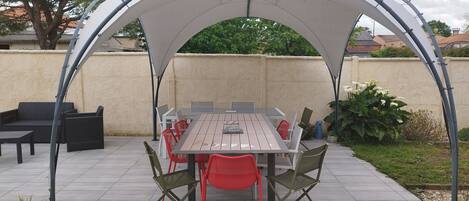 An outdoor patio with a covered dining area, including a table and chairs, surrounded by potted plants and a wall.