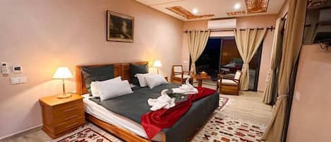A hotel room with a bed, two bedside tables, a nightstand, a painting on the wall, a large window with curtains, and a view of the outside.