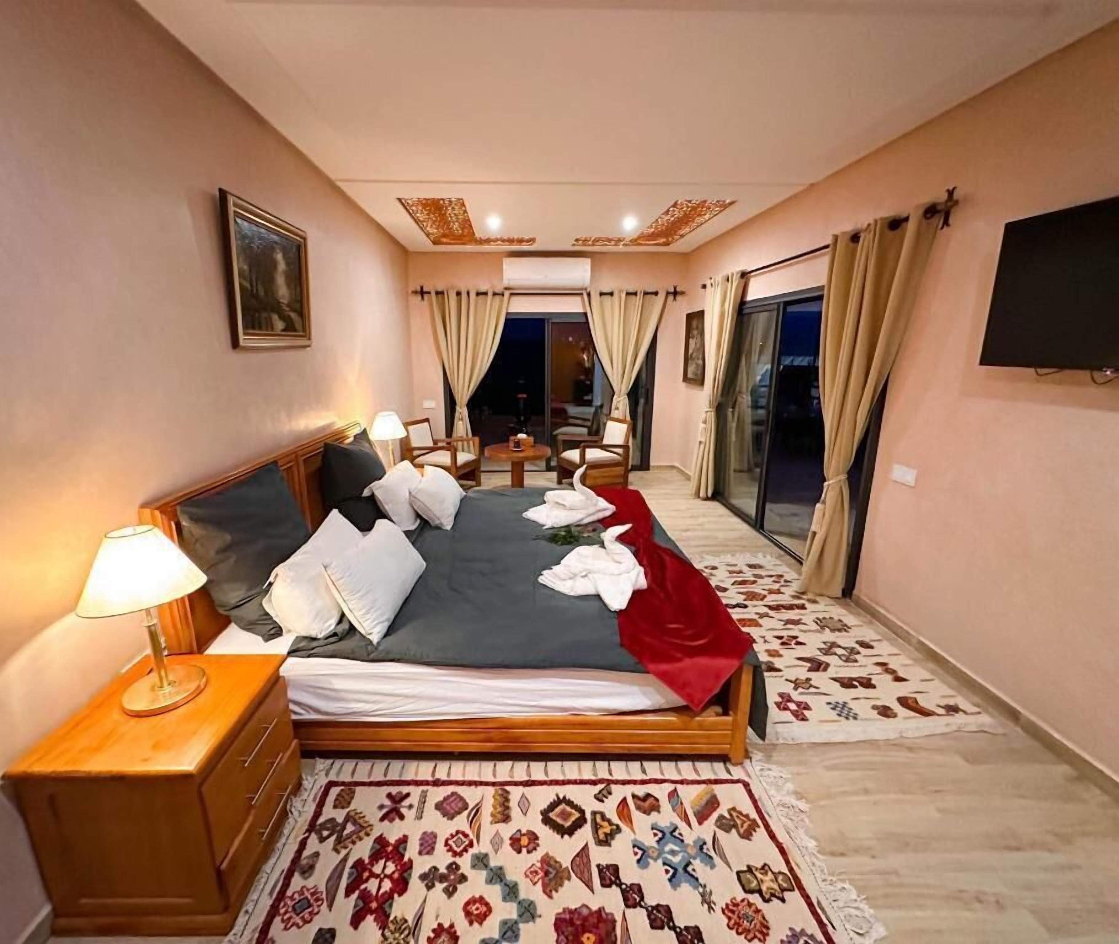 A hotel room with a large bed, a bedside table with a lamp, a TV mounted on the wall, and a view of the outside through a sliding glass door.
