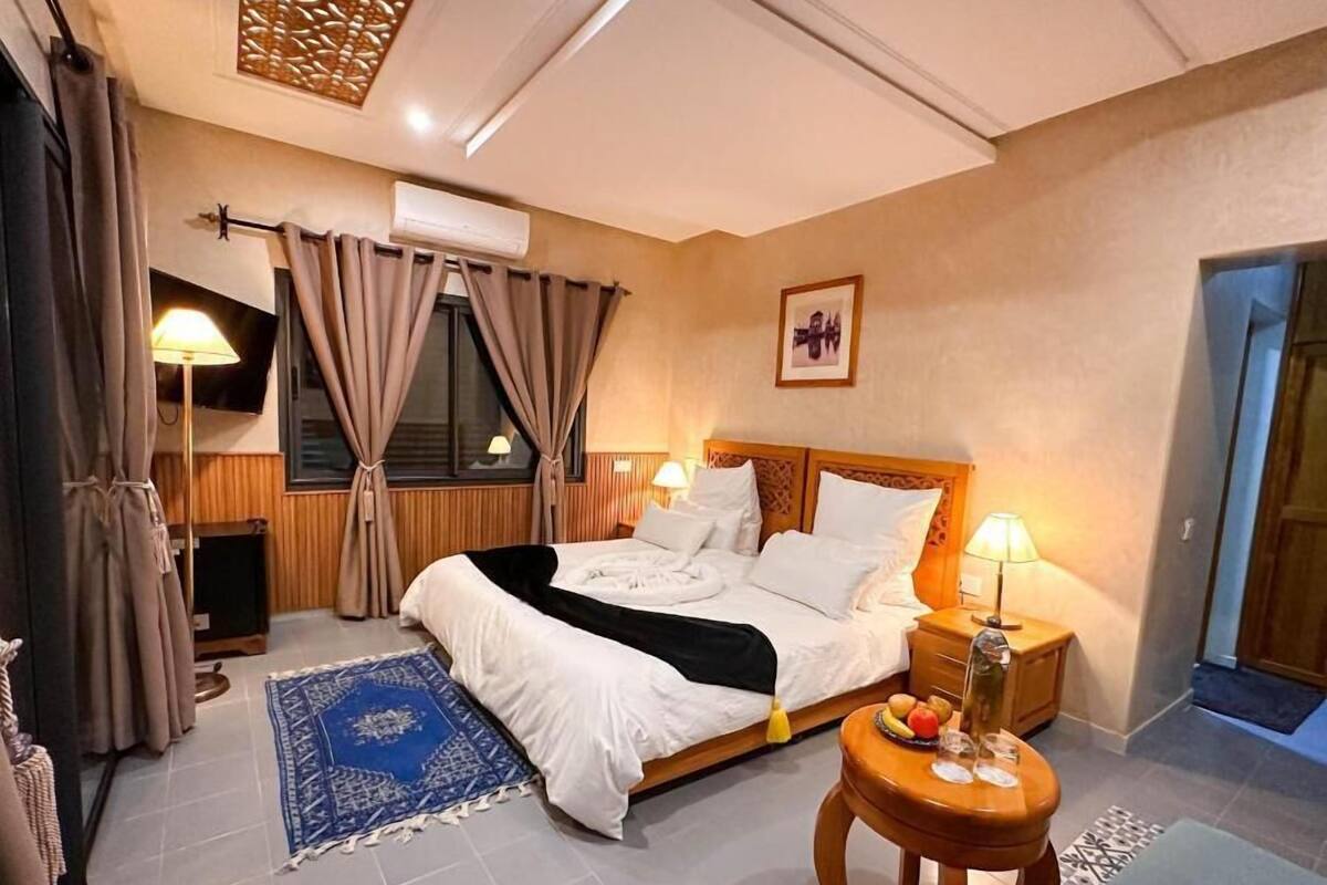 Ett hotellrum med en säng, sänglampor, ett litet bord med frukt, en blå matta och en luftkonditioneringsanläggning.